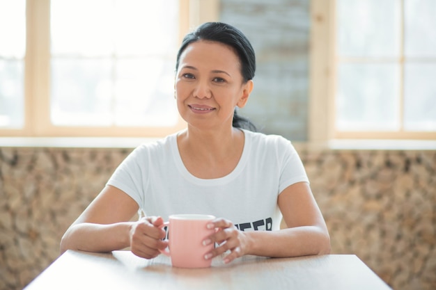 Coffee break. Happy glad female volunteer sitting at table and enjoying coffee while looking at camera