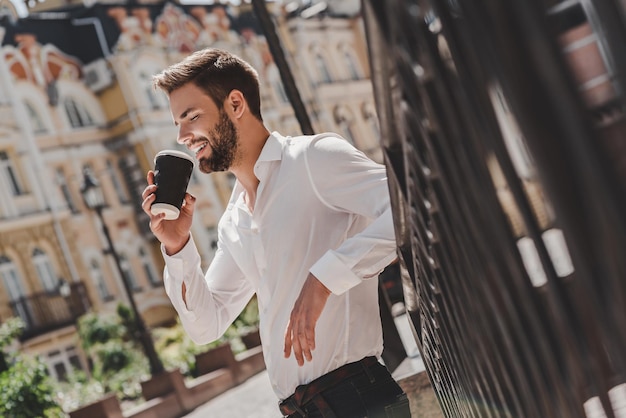 Ritratto di pausa caffè di uomo fiducioso dai capelli castani che tiene una tazza di caffè appoggiata sulle scale dopo