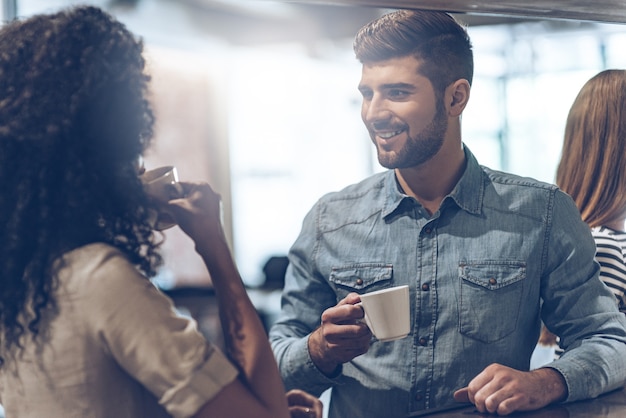 喜びでコーヒーブレイク。コーヒーカップを持って、バーのカウンターに立っている間、若い女性と何かを話し合う若いハンサムな男