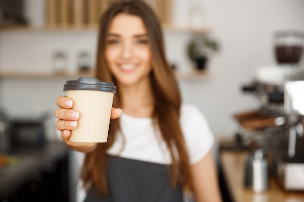 Coffee Business Concept - Beautiful Caucasian lady smiling at camera offers disposable take away hot coffee at the modern coffee shop