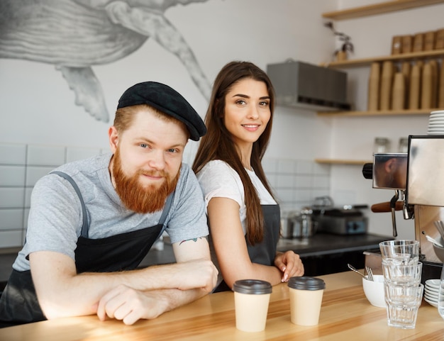 コーヒービジネスコンセプトモダンなコーヒーショップでコーヒーサービスを提供する準備ができているバークーターに立っている間エプロンでポジティブな若いひげを生やした男と美しい魅力的な女性バリスタカップル