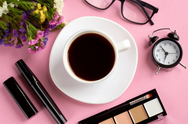 Coffee cup, alarm clock, flowers, mascara and glasses on pink surface.