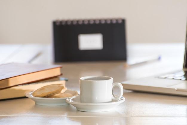 Coffee cup and in the background out of focus an agenda stacked books and a laptop computer