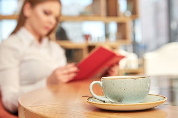 Foto tazza di caffè in primo piano, donna che legge un libro