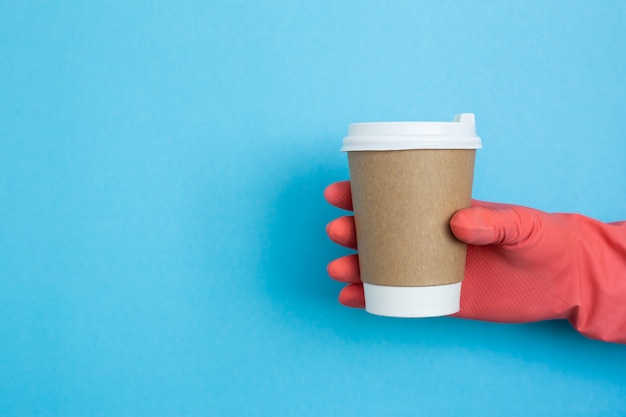 Фото Кофейная чашка в руке при розовые медицинские перчатки изолированные на голубой предпосылке. женская рука с бумажным стаканчиком. макет женской руки, держащей бумажный стаканчик кофе. копировать пространство коронавирус защита