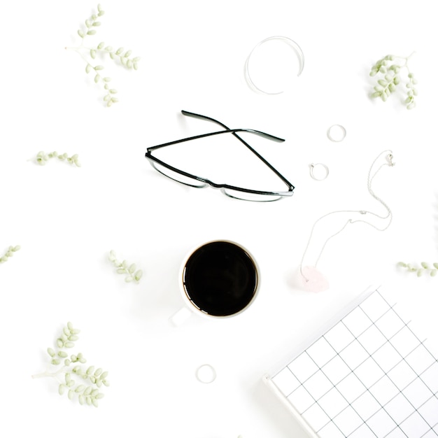 Coffee cup, notebook, glasses, green branches and female accessories on white surface