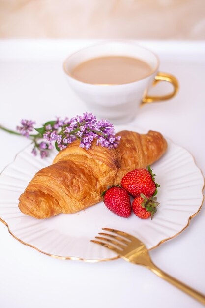 Coffee cup and sweet croissant with fresh strawberry on white table, decorated with flower