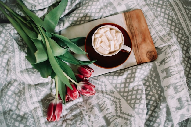 Coffee cup with marshmallows and red tulips in bed. Concept of holiday, birthday, Valentines Day, March 8