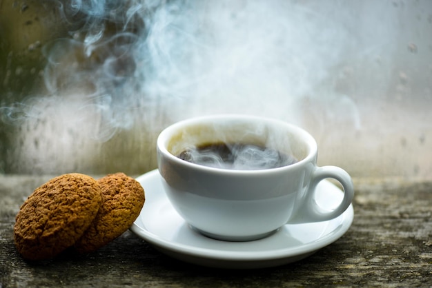 Coffee drink with oat cookies dessert enjoying coffee on rainy\
day coffee time on rainy day fresh brewed coffee in white cup or\
mug on windowsill wet glass window and cup of hot caffeine\
beverage