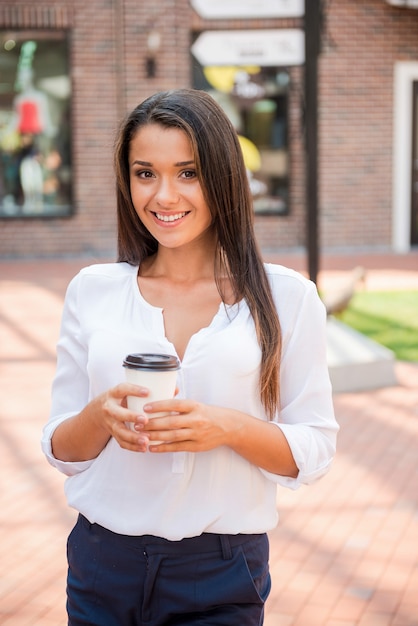 外出先でのコーヒー。コーヒーカップを持って、屋外に立って笑っている美しい若い女性