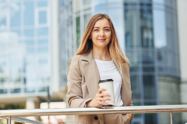 Coffee in hands Woman in formal wear standing outdoors in the city at daytime