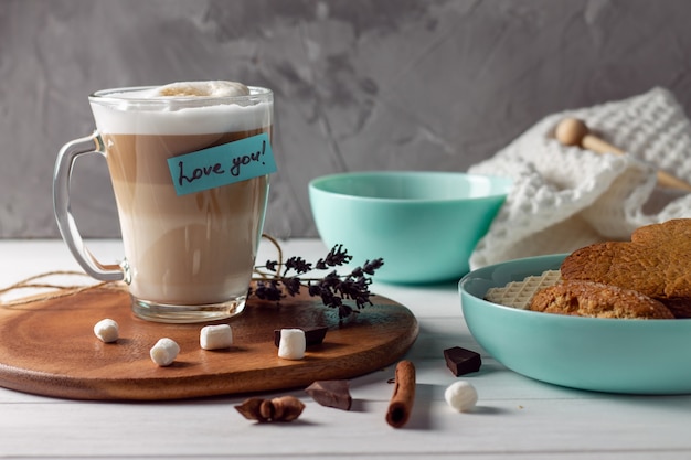 Coffee latte cup with Love you sticker on wooden tray with turquoise bowls with cookies and marshmallows on gray wall surface