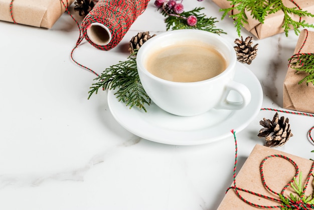Coffee latte mug with Christmas gift or present box wrapped in kraft paper, decorated with christmas tree branches, pine cones, red berries, on white marble table, copy space