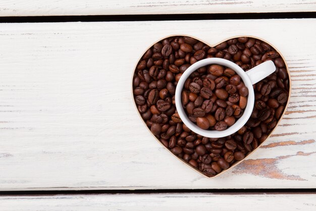 Coffee lover concept. Heart shape form filled with coffee beans. White planks on surface.