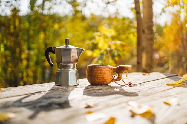 Coffee maker and wooden mug kuksa on a wooden table in the autumn forest.