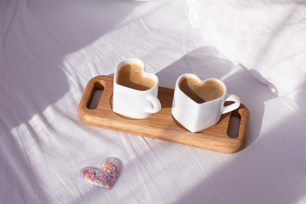 Coffee in mugs in the shape of a heart on a tray lie on a white bed