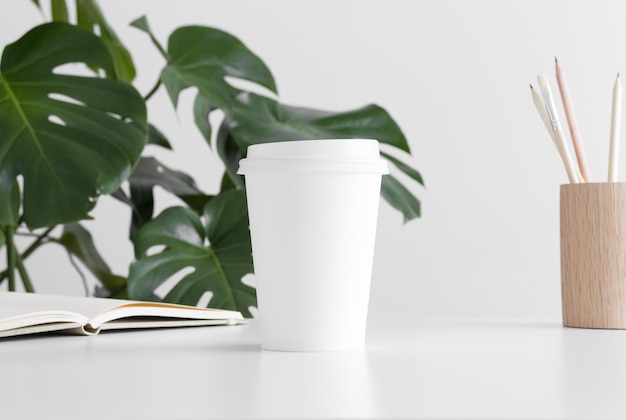 Coffee paper cup mockup with a notebook workspace accessories and a monstera plant on a white table