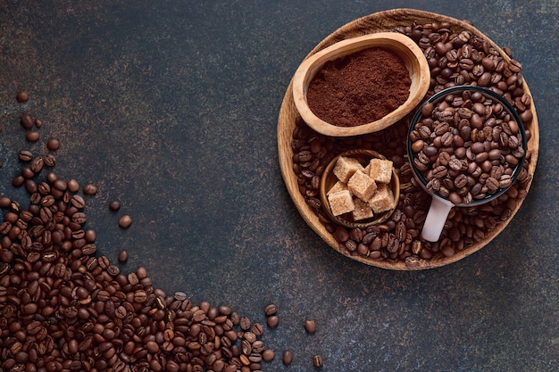 Chicchi di caffè tostati in tazza e sparsi nelle vicinanze, caffè macinato e zucchero di canna su un tavolo marrone vista dall'alto con spazio per copiare il testo.