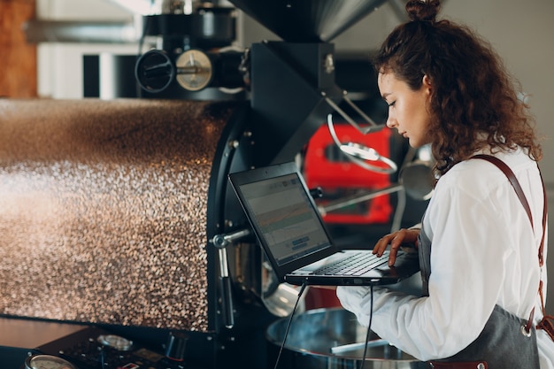 Macchina della torrefazione del caffè e donna del barista con il computer portatile al processo di torrefazione del caffè