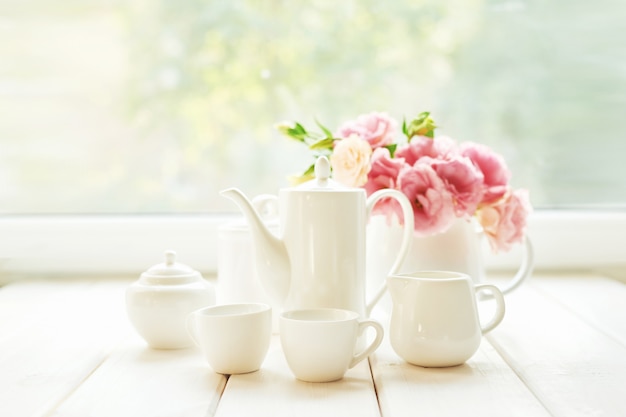 Photo coffee set next to a vase of flowers on a table against  a window