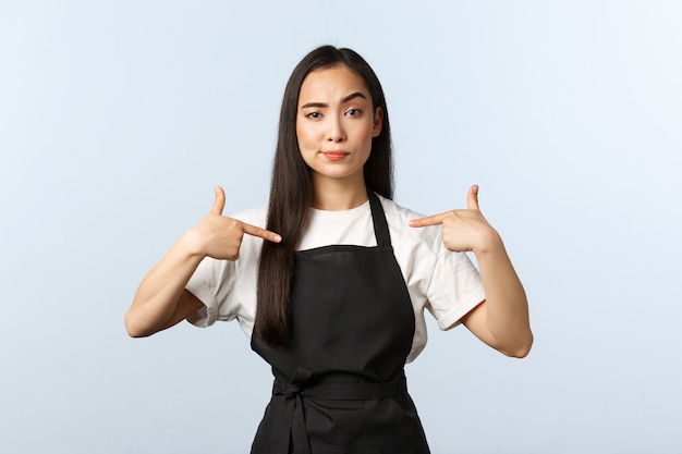 Coffee shop, small business and startup concept. Confident young asian woman, professional barista in black apron look serious and pointing herself, show-off as best cafe in town