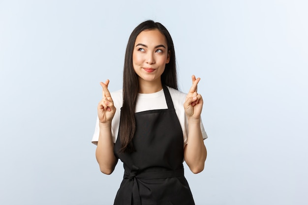 Coffee shop, small business and startup concept. Cute hopeful asian barista, waitress or cafe staff in black apron making wish, smile and look away imaging dream come true as cross fingers good luck.