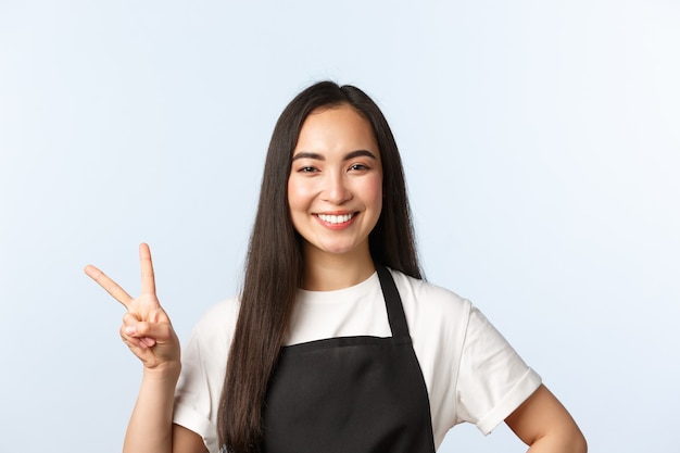 커피숍, 중소기업 및 시작 개념. 친절하고 귀여운 아시아 여성 직원, 검은색 앞치마를 입은 카페 직원은 평화 사인을 보여주고 고객에게 미소를 짓고 긍정적인 태도를 표현합니다.