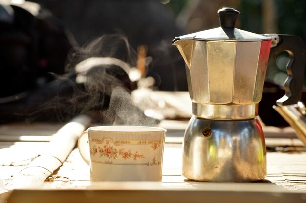 Coffee steamer and a cup with steam on bamboo table in a morning