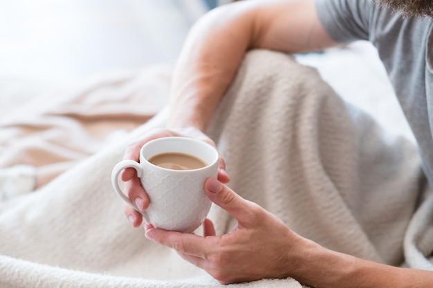 コーヒータイムホーム居心地のよさの楽しみ休日の男ベッドの男手にラテのカップ