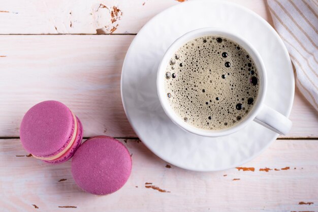 Coffee in a white cup and two pink macaroons on a light wooden background top view