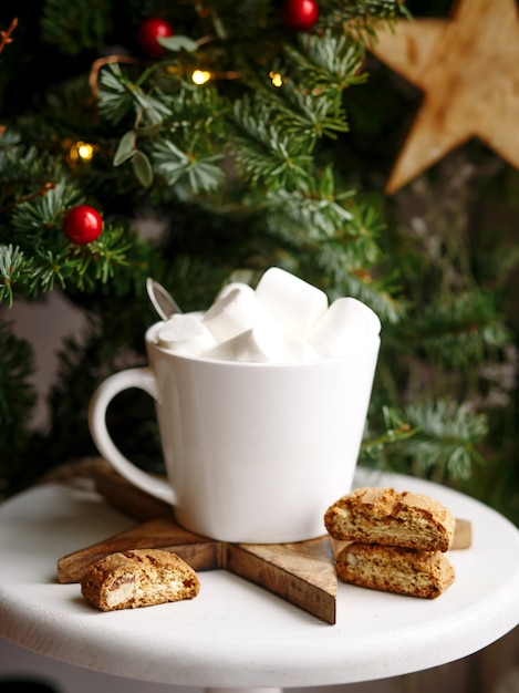 Coffee in a white cup with marshmallows. Morning festive coffee with traditional Italian cantuccini almond cookies. A cup of coffee on a background of green fir branches on a white stand.