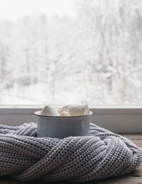 Coffee with marshmallows and a cozy grey sweater on vintage windowsill against snow landscape from outside. Soft focus. Relaxing winter day at home with traditional winter hot drink. Minimalist style.