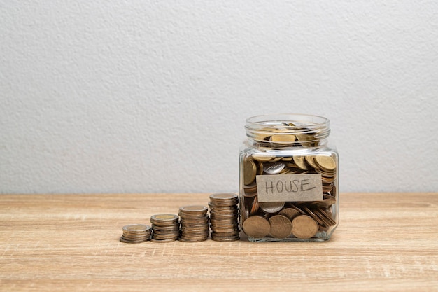 Coin in a glass jar with money stack on the brown table for saving to buy house, financial concept