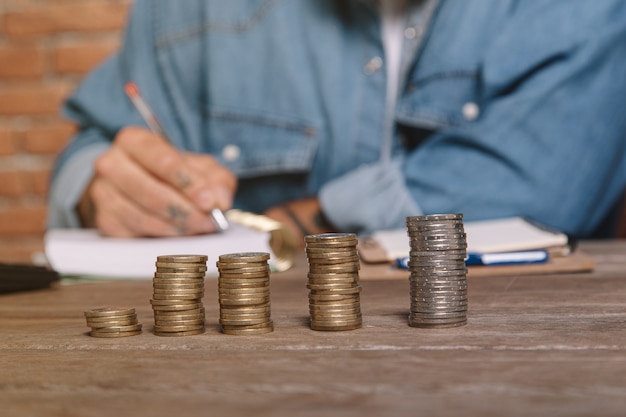 Coins stacked in the foreground and man writing down expenses in a notebook to calculate the concept of saving money for household accounting.