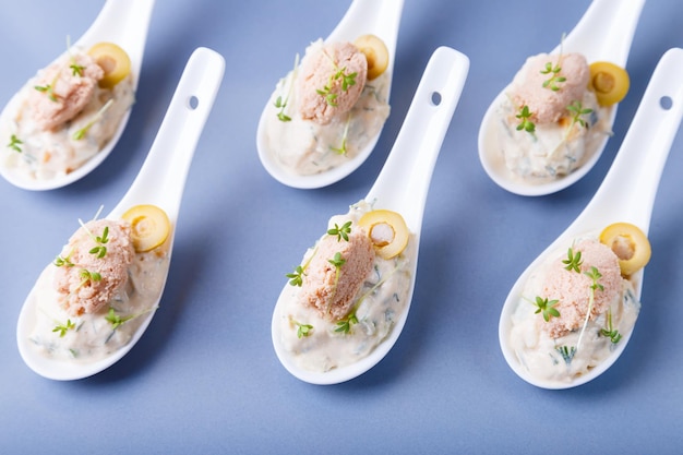 Photo cold appetizer made from cod liver, cod caviar, olives, cucumber and microgreens on serving spoons. traditional cold dish. close-up, blue plate, black background.