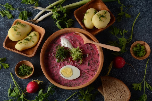 Cold beetroot soup with vegetables on dark grey table. Traditional Ukrainian Russian Polish beet soup. Cold summer soup served with boiled potatoes and eggs, and whole grain bread in a wooden bowl