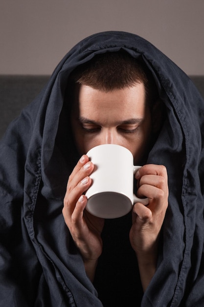 Cold and flu Portrait of a sick young guy drinking tea in bed sick caucasian sitting with a cup of hot drink in his hands with a fever not feeling well at home closeup