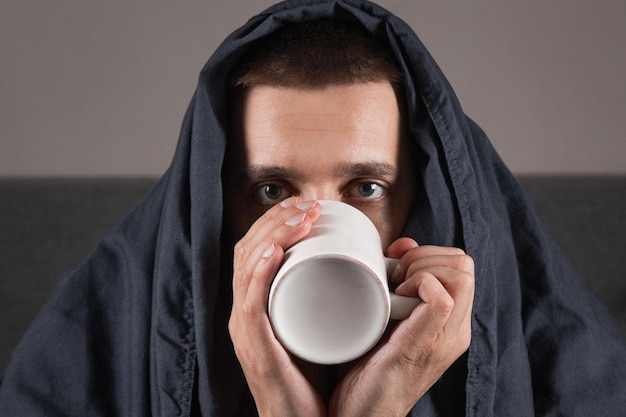 Простуда и грипп Портрет больного молодого парня, пьющего чай в постели, больного кавказца, сидящего с чашкой горячего напитка в руках с лихорадкой, плохо себя чувствующего дома крупным планом