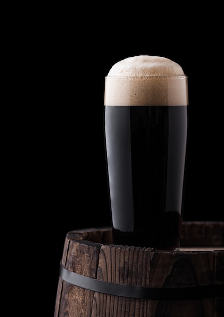 Cold glass of dark stout beer on wooden barrel on black background