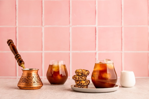 Photo cold iced coffee in a glass, served with oatmeal cookies. a cezve and white milk jug. warm light and bright image, pink tile backdrop