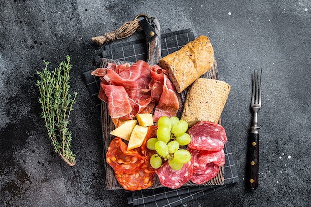 ハムプレート、豚肉-パンとブドウを添えた木の板に伝統的なスペインのタパス。黒の背景。上面図。