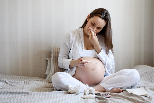 Cold pregnant woman sitting with thermometer in bed with tablets and wipes