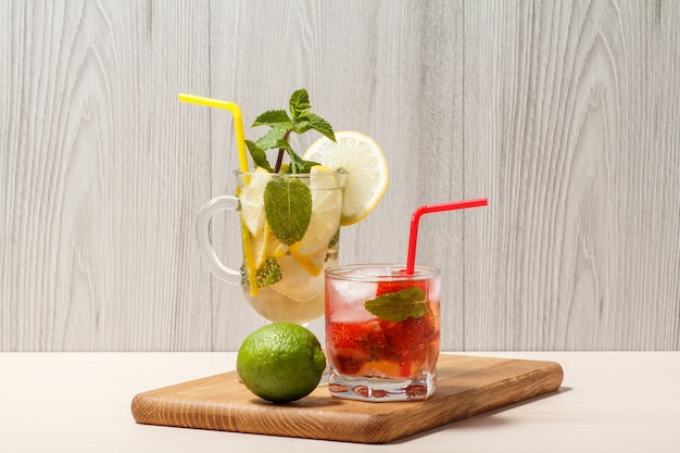 Cold refreshing summer lemonade mojito with lemon slices and mint in a tall glass and strawberry lemonade with mint in a short one on wooden cutting board