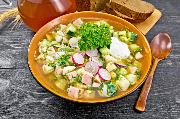 Cold soup okroshka from sausage potatoes eggs radish cucumber greens and kvass in a bowl bread and jug with drink on the background of wooden board