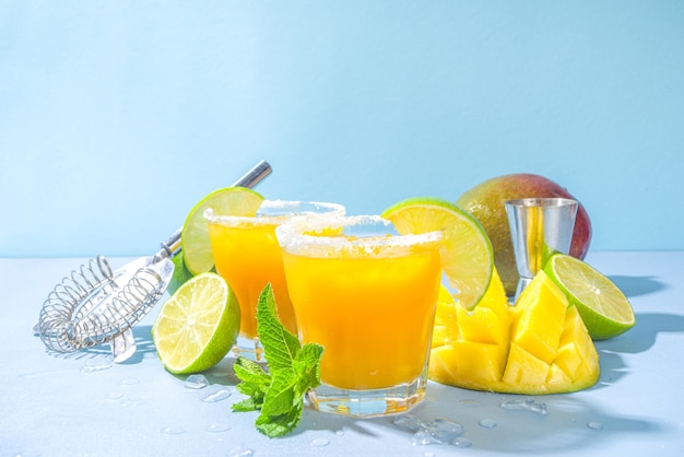 Cold summer cocktail, mango margaritas with tequila, salt lime slices, crushed ice and mint. Seasonal refreshing drink, on bright blue sun lighted background