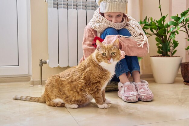 Cold winter autumn season girl child in scarf and hat is warming herself near heating radiator with cat