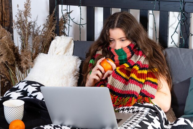 Photo a cold young woman with a warm scarf around her neck is sitting in bed and working with a laptop