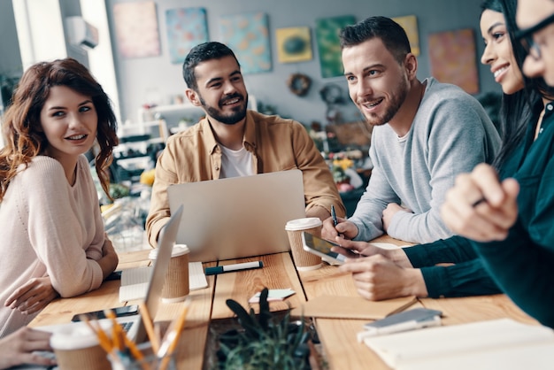 Collaboration. Group of young modern people in smart casual wear discussing something and smiling while working in the creative office