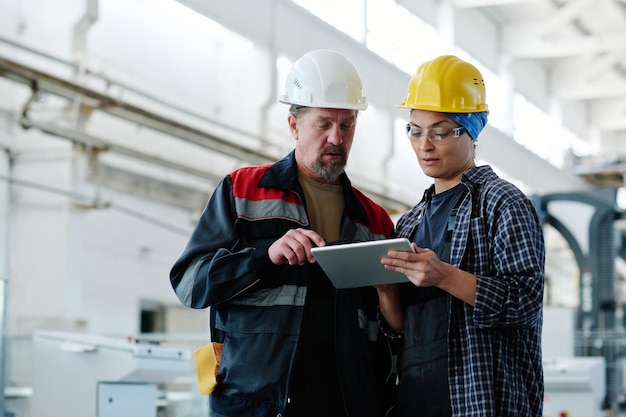 Colleagues in hardhats discussing project on digital tablet together during their work in factory