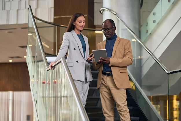 Colleagues standing on stairs in the hall and discussing online presentation on tablet pc